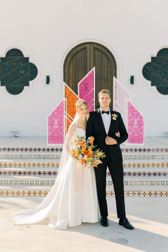 Bride and groom standing in front of the La Palmilla Chapel, showcasing vibrant, colorful wedding florals in a garden-style design.