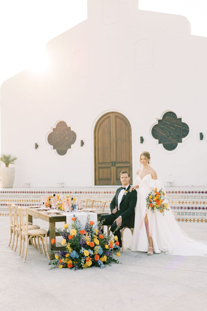 Head table with vibrant floral arrangements, featuring bright flowers on top and a ground floral at the end, with the bride and groom standing nearby.