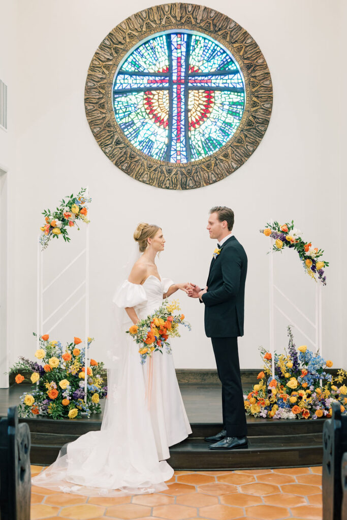 Bride and groom standing at the altar, looking at each other and embracing the moment, surrounded by vibrant, colorful wedding florals.