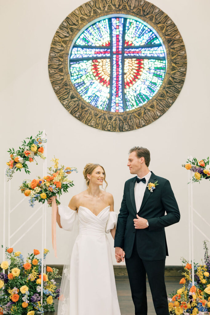 Here’s an SEO-friendly title and alt text for that image:
Bride and groom standing at the altar with vibrant floral arrangements behind them, featuring bright, colorful flowers in a garden-style design.